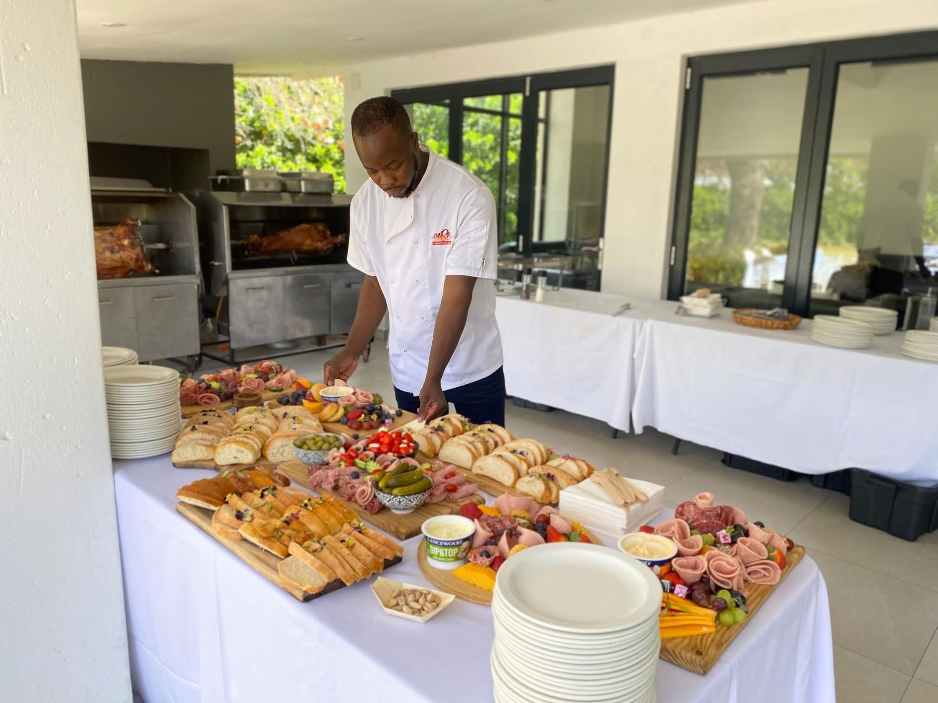 Chef arranging a large charcuterie and sandwich buffet on a table outdoors.
