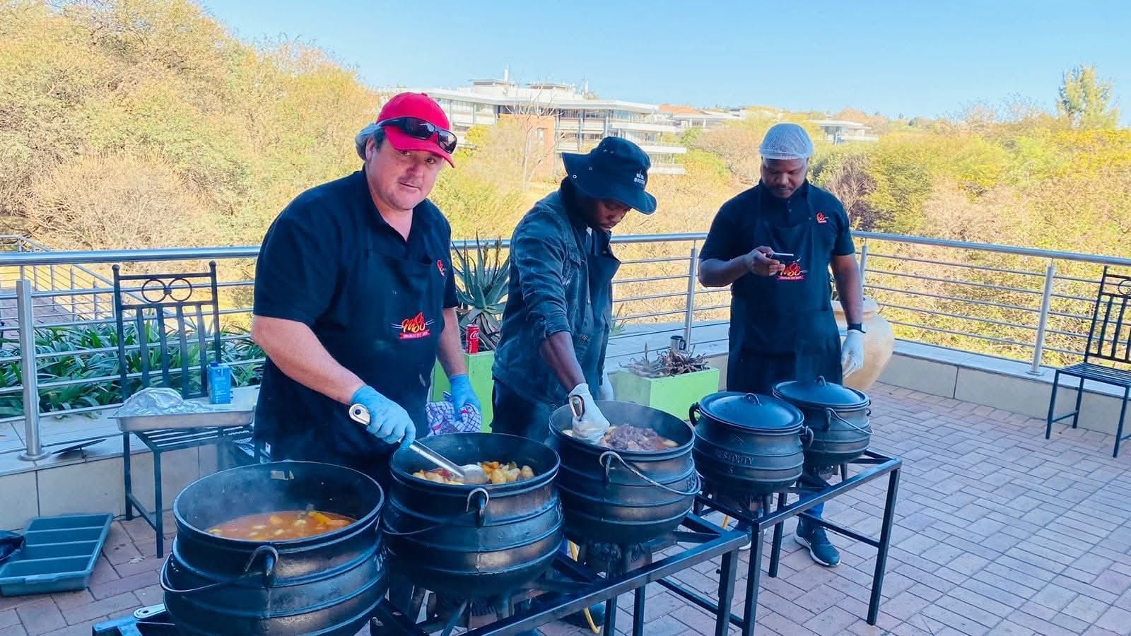 Three people cook in cast iron pots outdoors. One stirs, one adds food, and one holds a phone.