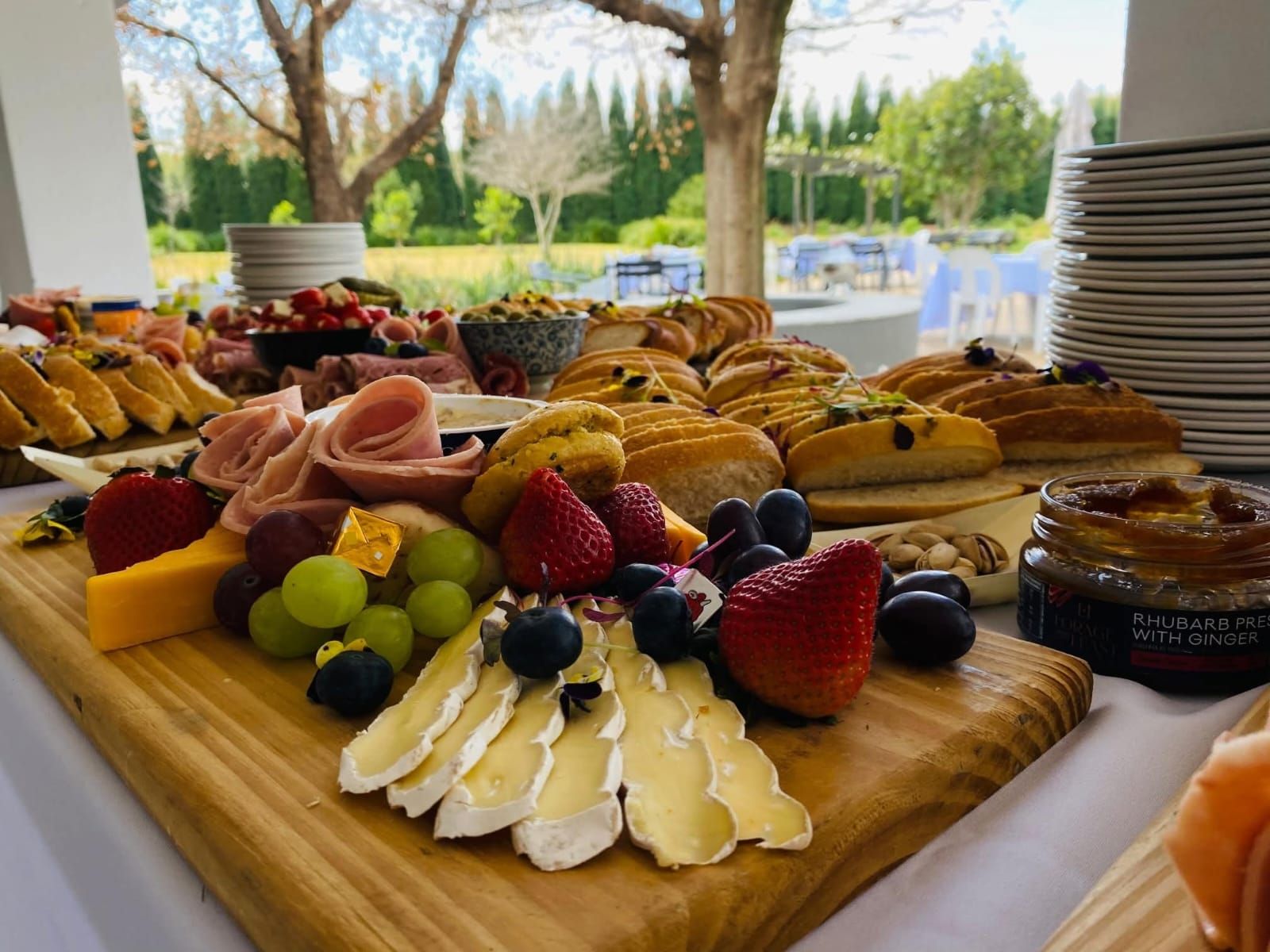 Charcuterie board with fruit, cheese, pastries, and meats set outside near a garden.