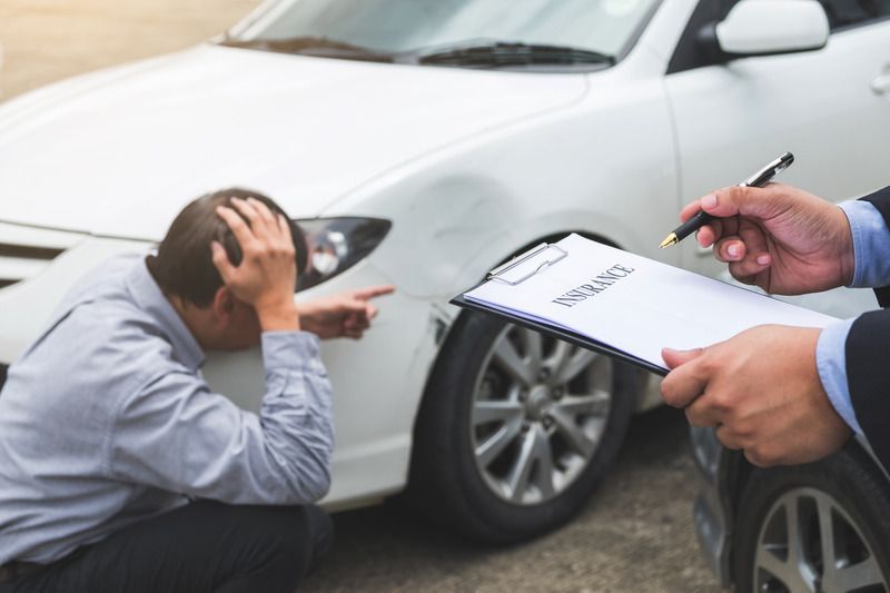 Man distraught, pointing at car damage. Another man in suit holding clipboard, taking notes. White car with dent.