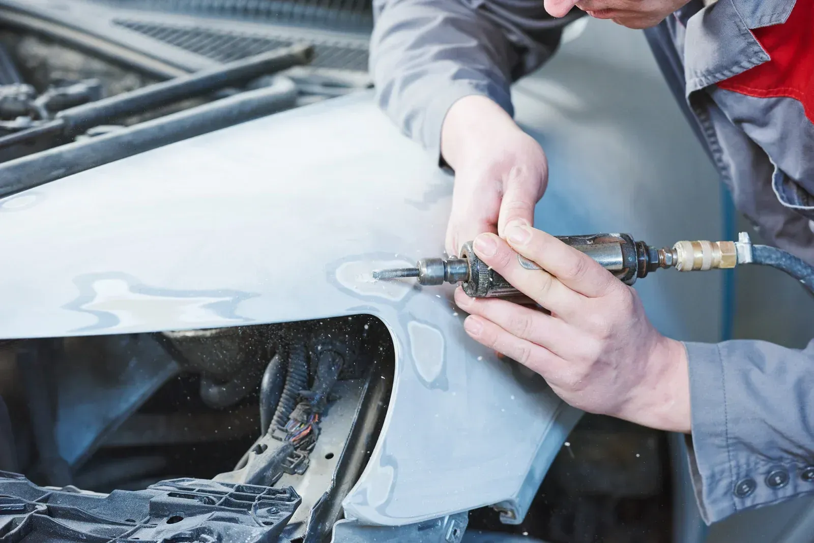 Mechanic using a pneumatic sander to repair a dent on a gray car fender.