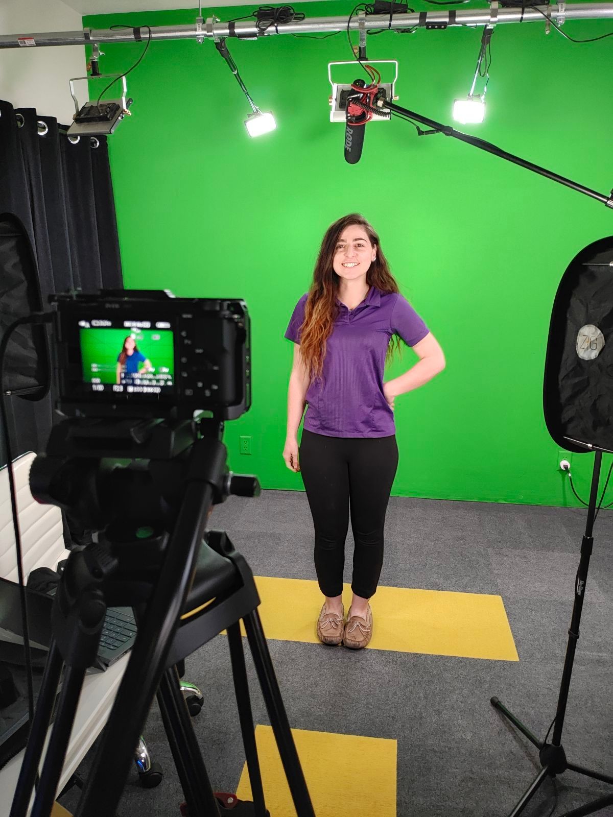 A woman in a purple shirt is standing in front of a green screen