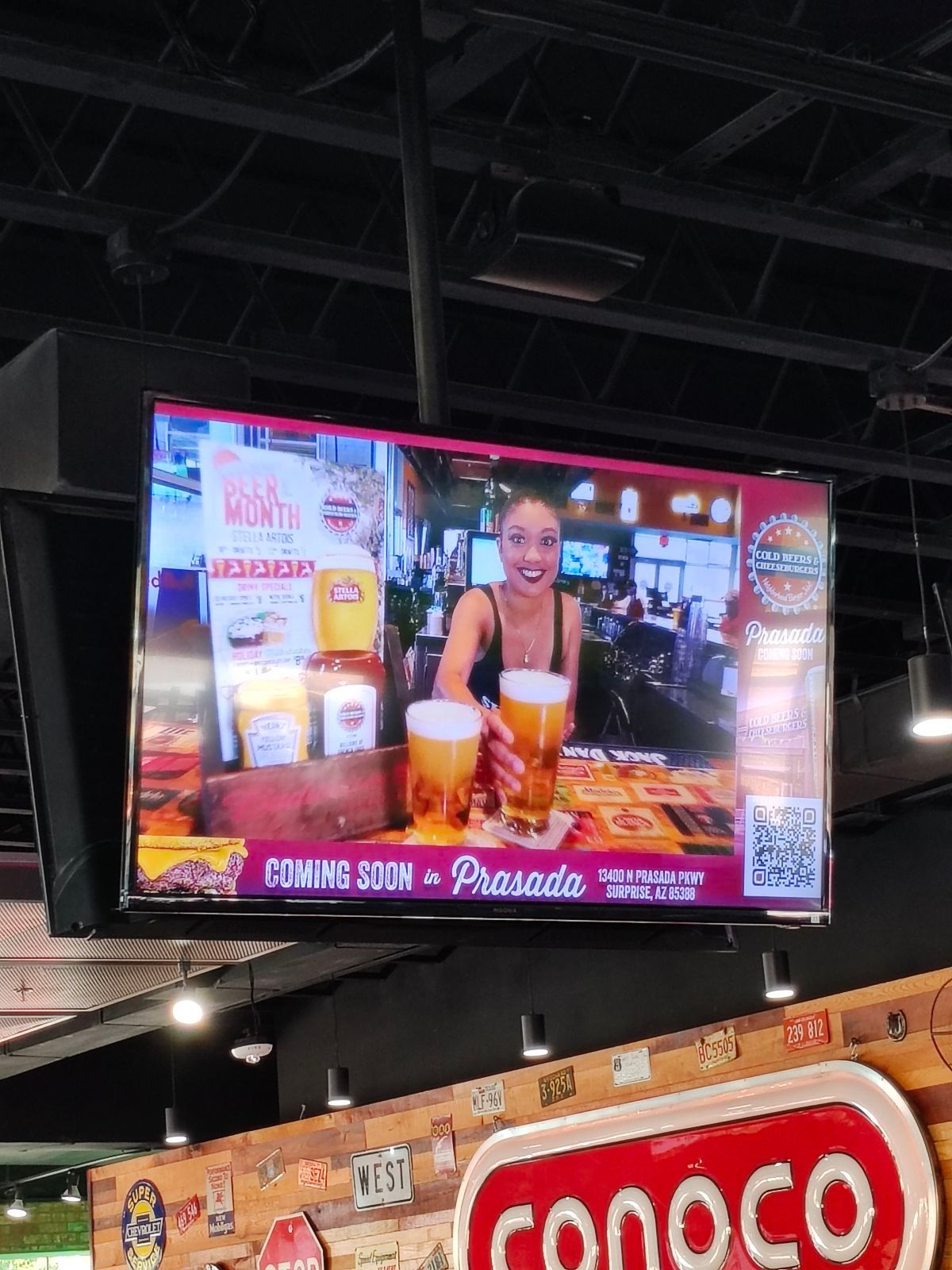 A tv screen hanging from the ceiling of a restaurant shows a woman holding a glass of beer.