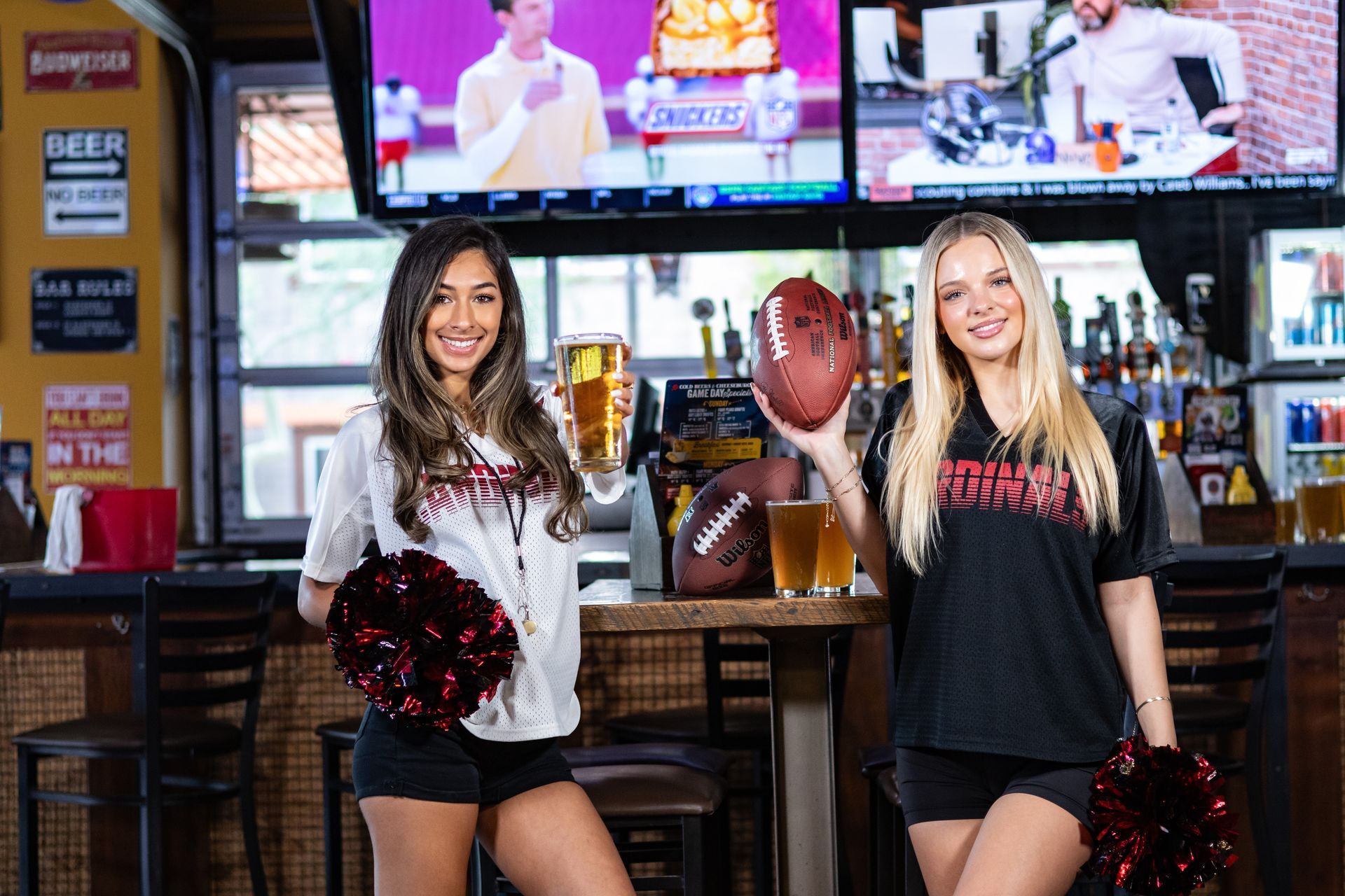 Two cheerleader models are standing next to each other in a bar holding beer and a football.