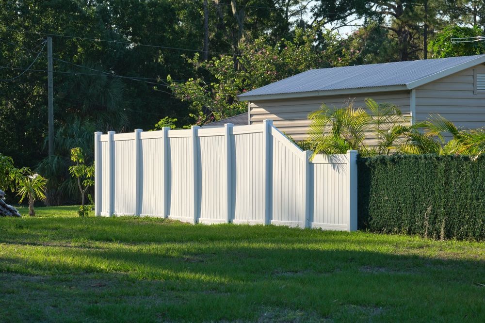White vinyl fence partially bordering a lawn, with a shed and foliage in the background under a bright sky.
