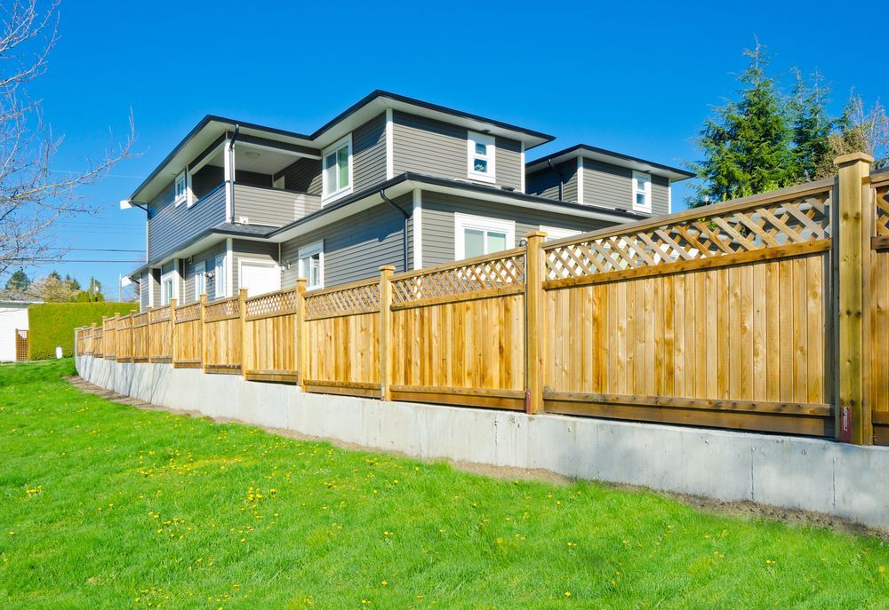 Two-story gray house with a wooden fence and a green lawn under a clear blue sky.