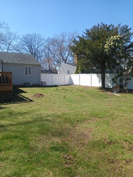 A backyard with a white fence, grass, and a tree.  A house is on the left. Sunny day.