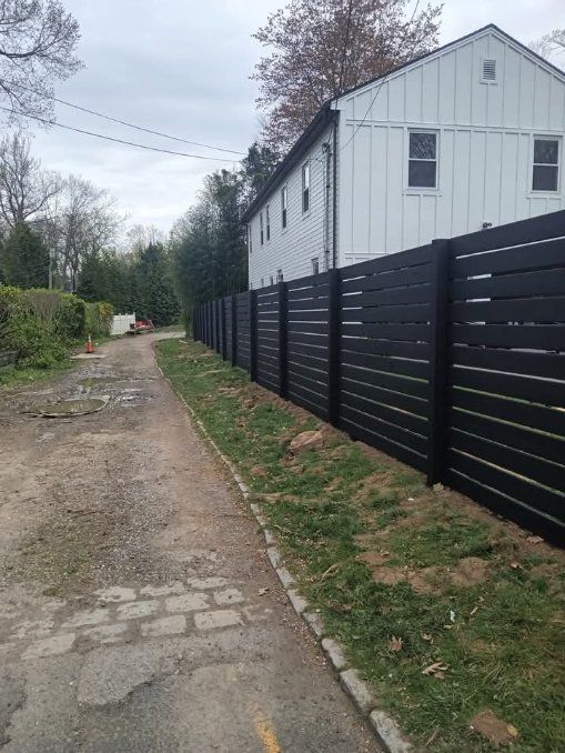 Black horizontal slat fence alongside a gravel driveway, with a two-story white house in the background.