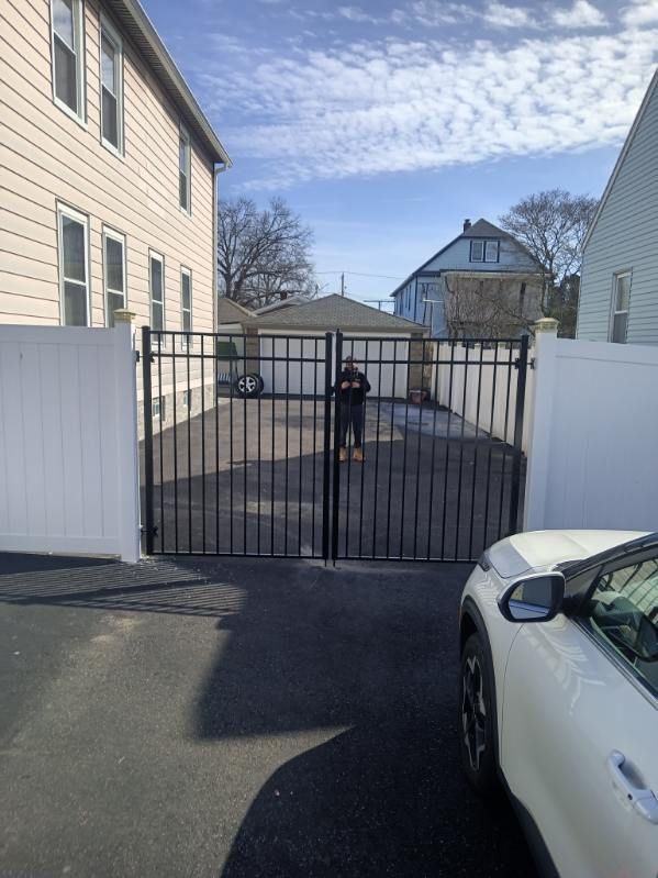 Black iron gate opening to a paved driveway. White fences flank the sides. A person stands near the gate.