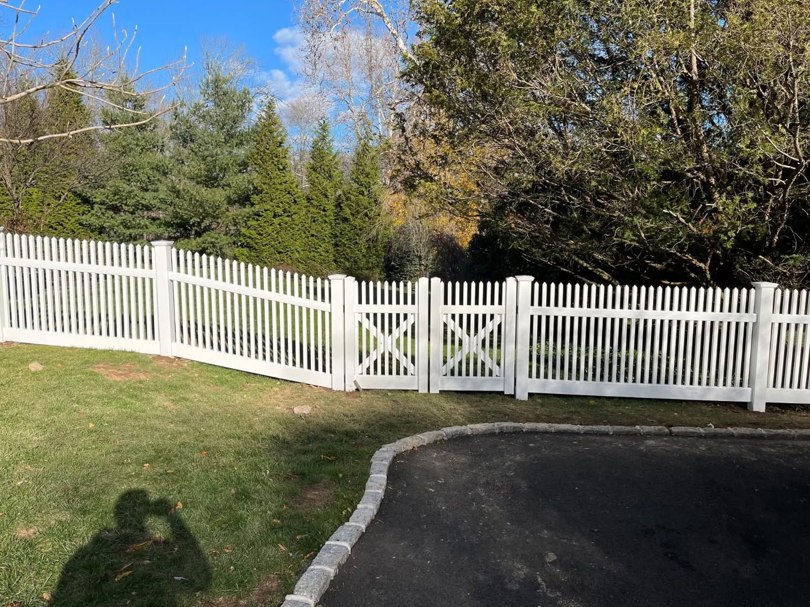 White picket fence with gate enclosing green lawn and trees, next to black asphalt driveway.