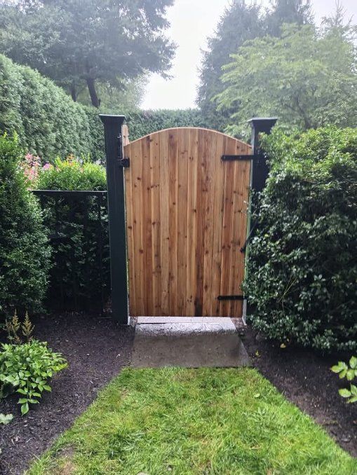 Wooden garden gate, arched top, flanked by dark pillars and lush green hedges. Path of grass and stone leads to it.