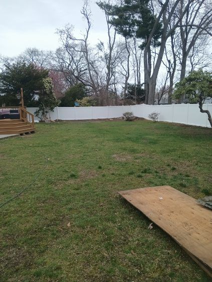 Backyard with green grass, white fence, and trees. A wooden board lies in the foreground.