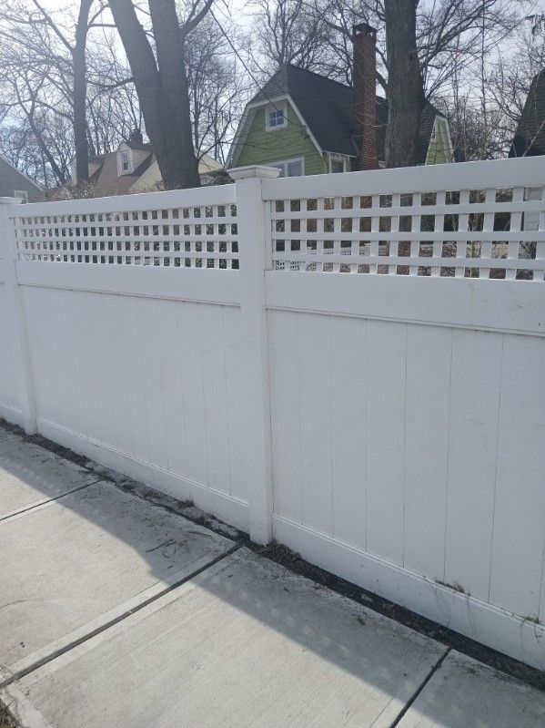 White vinyl fence with lattice top along a sidewalk, in front of trees and houses on a sunny day.