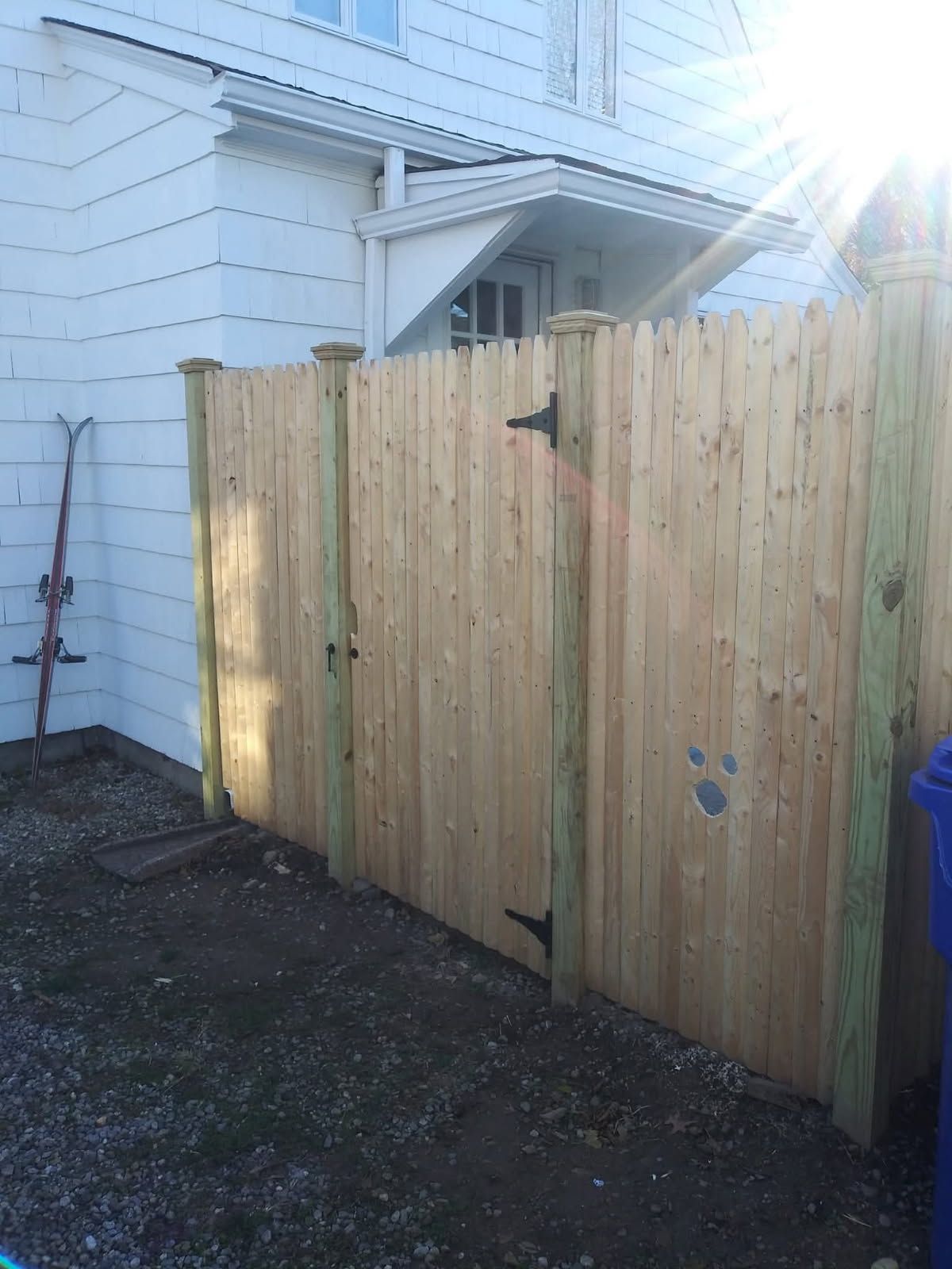 Wooden fence with gate, set against a white building. Sun shining.