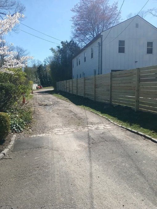 Paved driveway lined with a new wooden fence, leading towards a white building with windows on a sunny day.