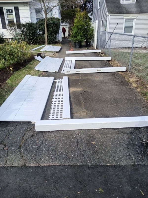 White fence components laid out on a driveway, preparing for fence assembly. A person walks in the background.