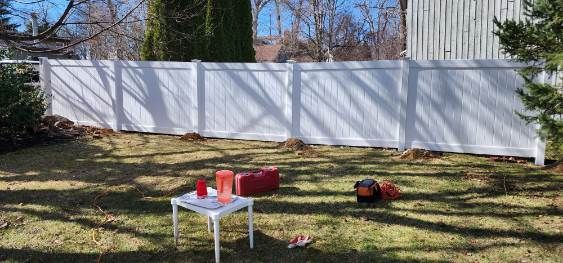 Backyard scene with a white picket fence. A small table holds drinks, and a lawnmower sits on the grass.