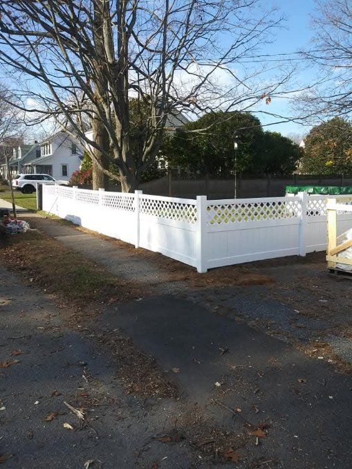 White lattice fence surrounds a driveway, next to a house with a bare tree in the background.