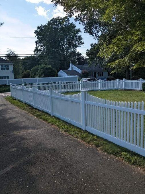 White three-rail fence alongside a paved driveway and green lawn, with trees in the background.