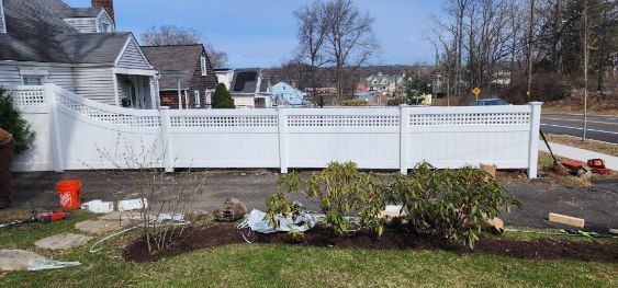 A white fence bordering a yard with landscaping, a driveway, and houses in the background. The sky is blue and the day is sunny.