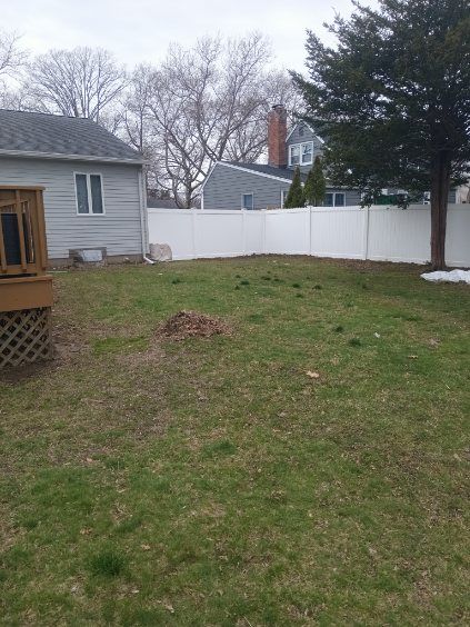 Backyard with a white fence, a house on the left, a large tree on the right, and patches of grass.
