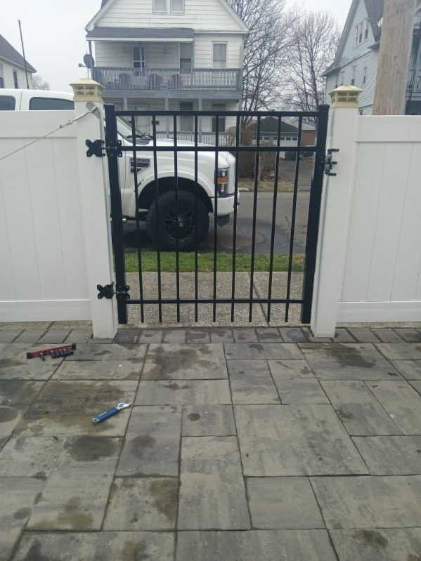 Black metal gate within a white fence on a stone patio, with a large truck visible behind it.
