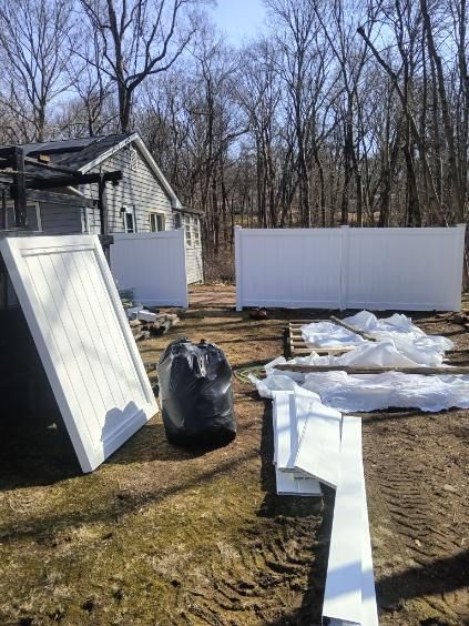 White vinyl fence being installed outdoors, with a black trash bag and construction materials nearby. A small house is visible in the background.