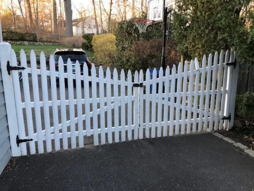 White picket fence with gate in front of a driveway, black hinges and latch.