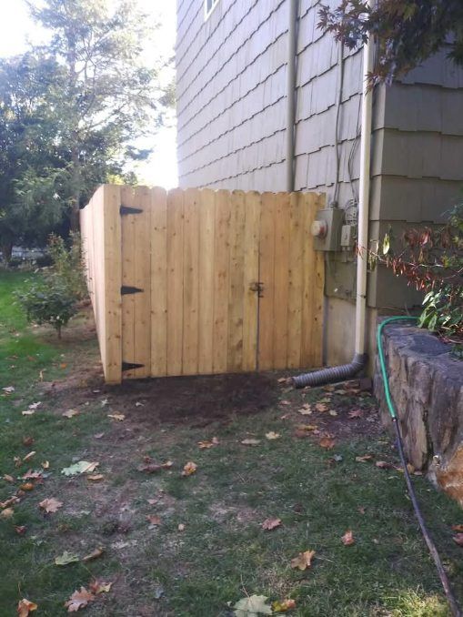 Wooden fence next to a house, enclosing utilities. Brown grass and fallen leaves in the yard.