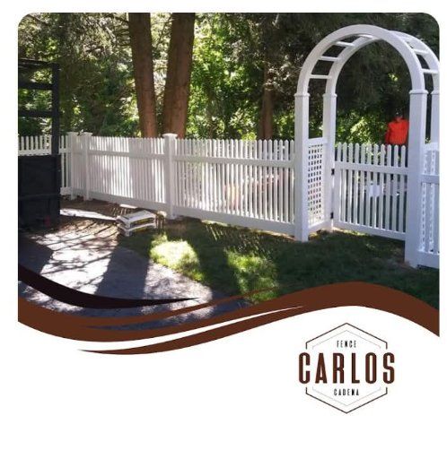 White picket fence with arched gate in a backyard setting, near trees. Logo in the lower right corner.