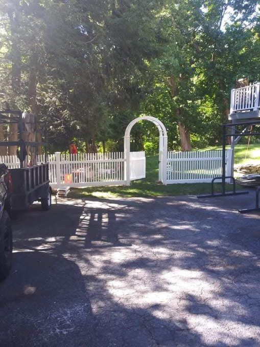 White picket fence with an archway, trees in the background, asphalt foreground.