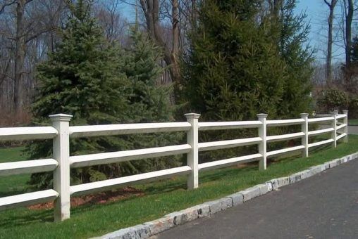 White three-rail fence alongside a grassy area next to a paved road, trees in the background.