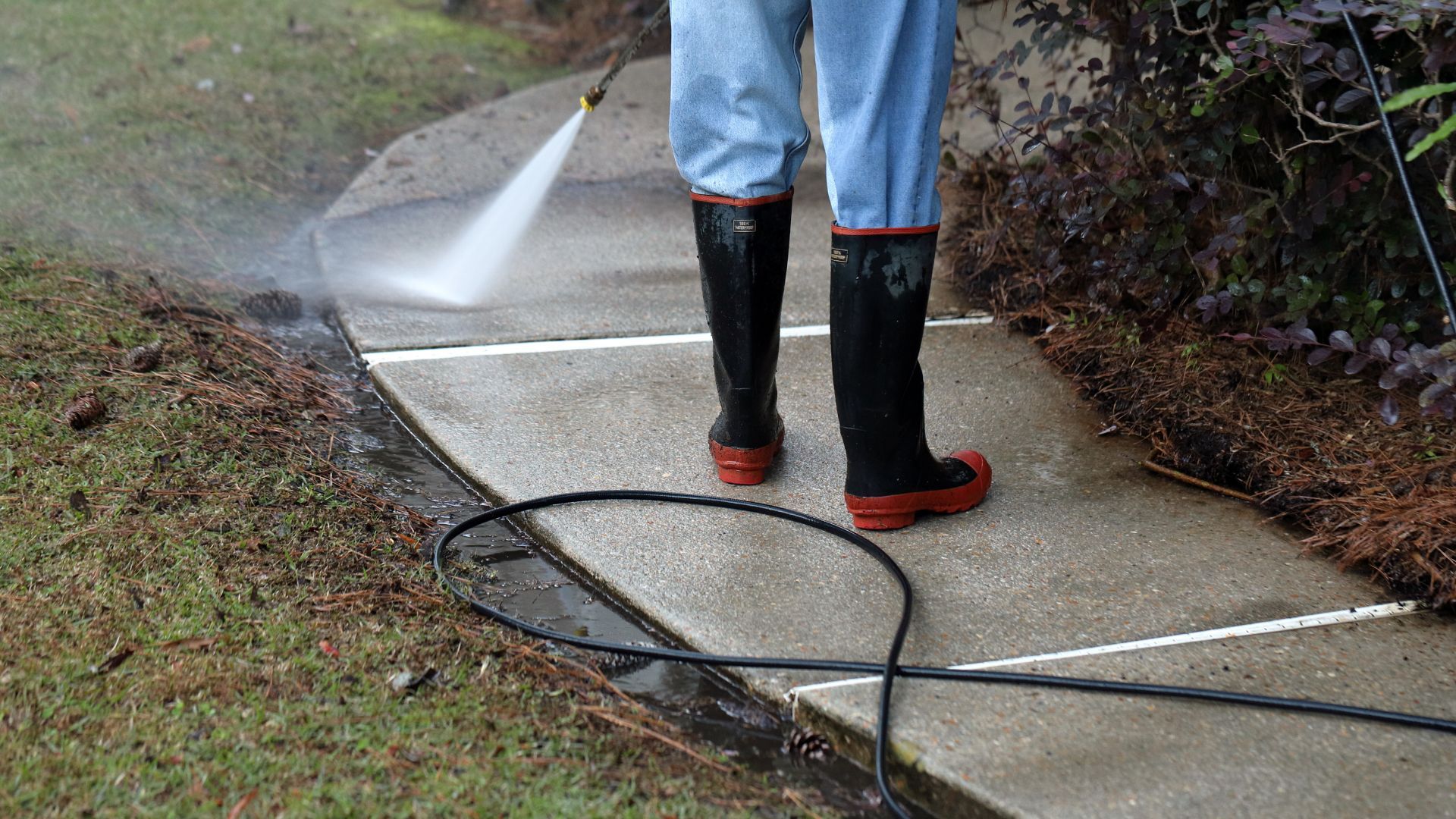 Person in boots pressure washing a concrete sidewalk.