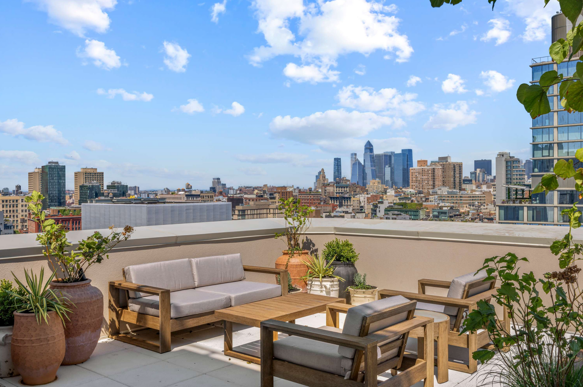 Rooftop patio with wooden furniture, city skyline view under blue sky with clouds.
