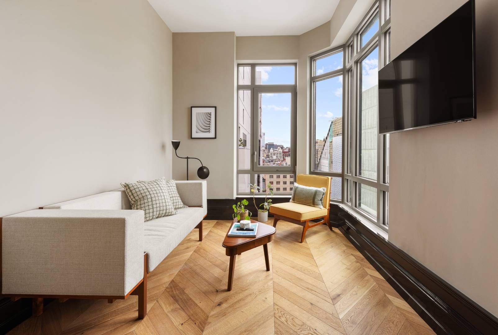 Living room with wooden floors, beige sofa, yellow chair, and large windows with city views.