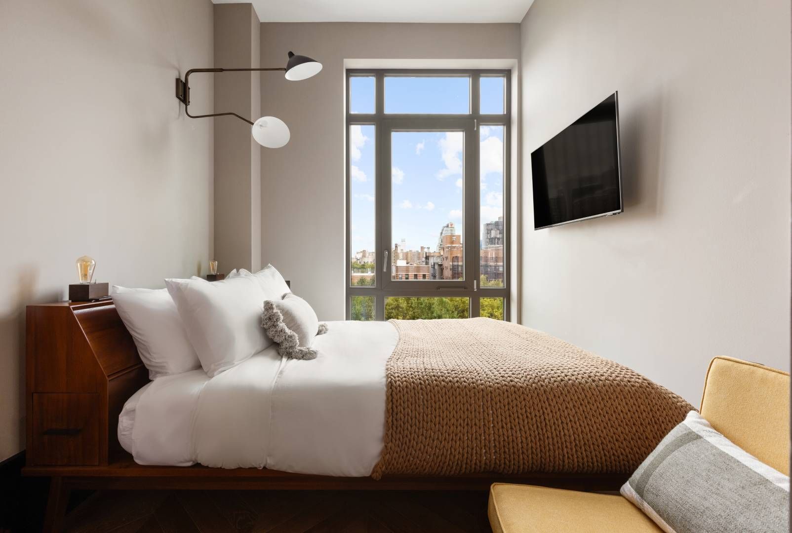 Bedroom with bed, window, TV, and a chair. Gray walls, wooden bed frame, tan blanket, and city view.