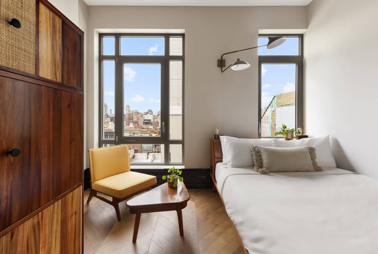 Small bedroom with a bed, chair, and wooden cabinet. Window view of city buildings.