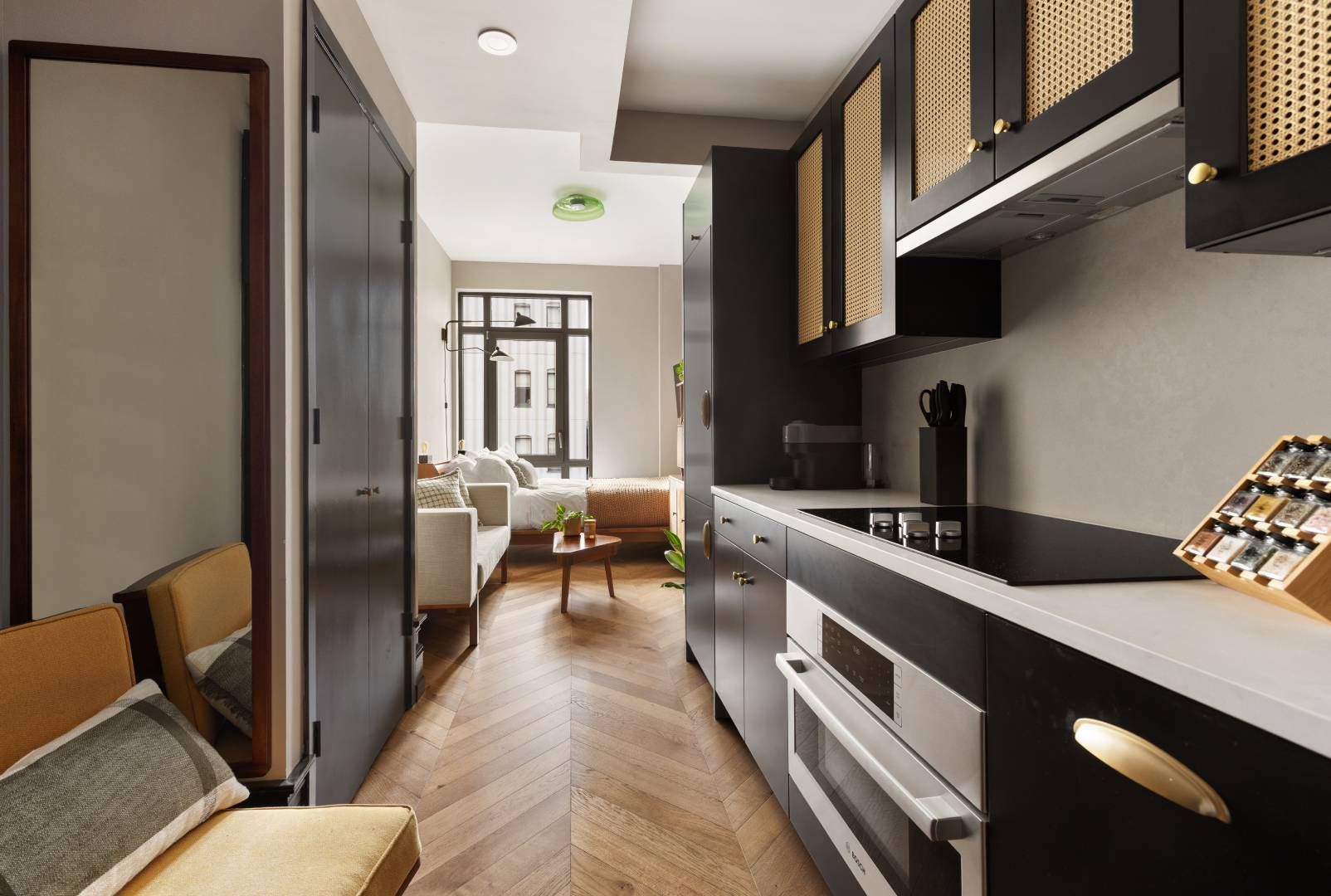 Narrow modern kitchen with dark cabinets, oven, and cooktop, leading to a living area.