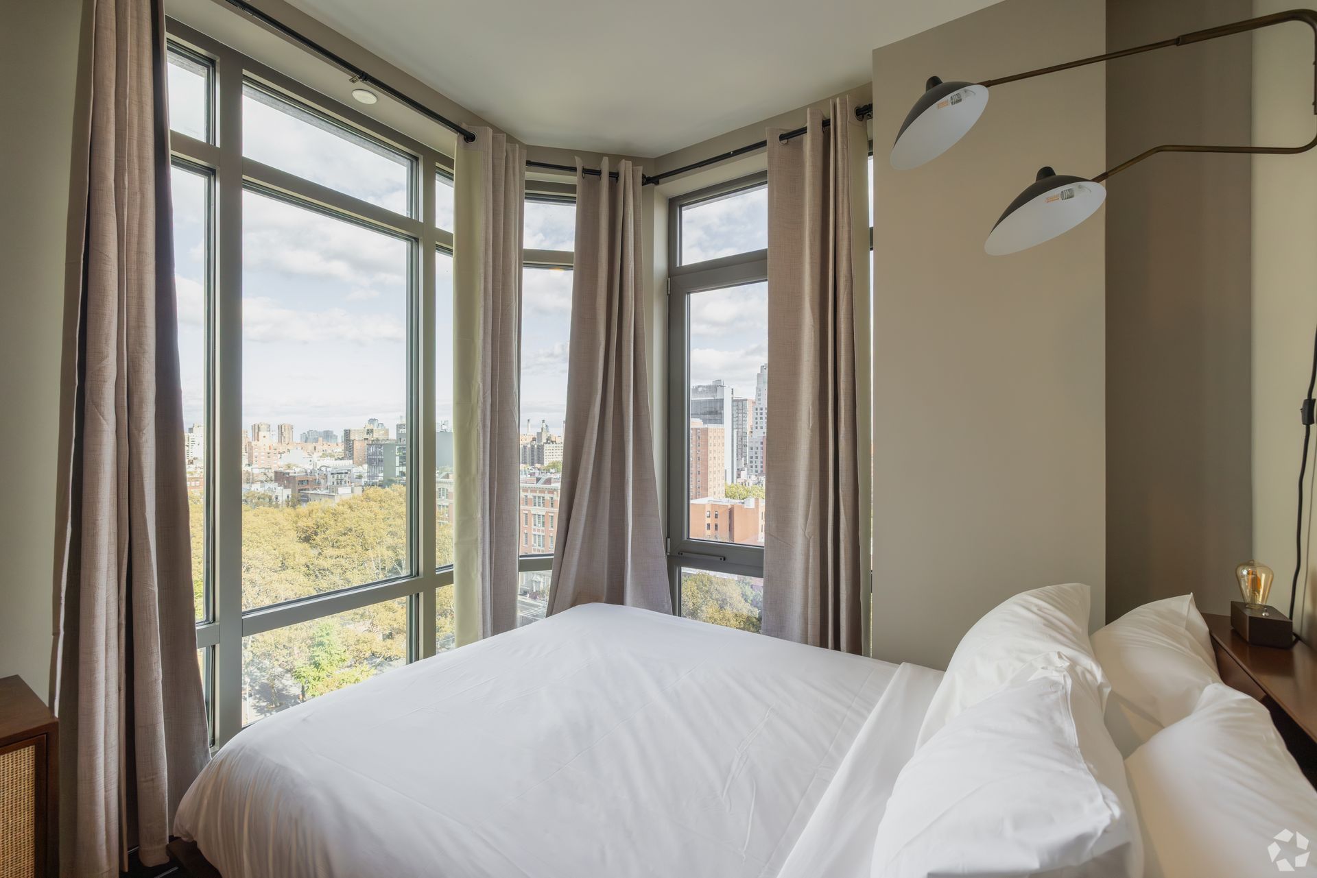 Bedroom with large windows, city view, bed with white bedding, gray curtains, and modern lighting.