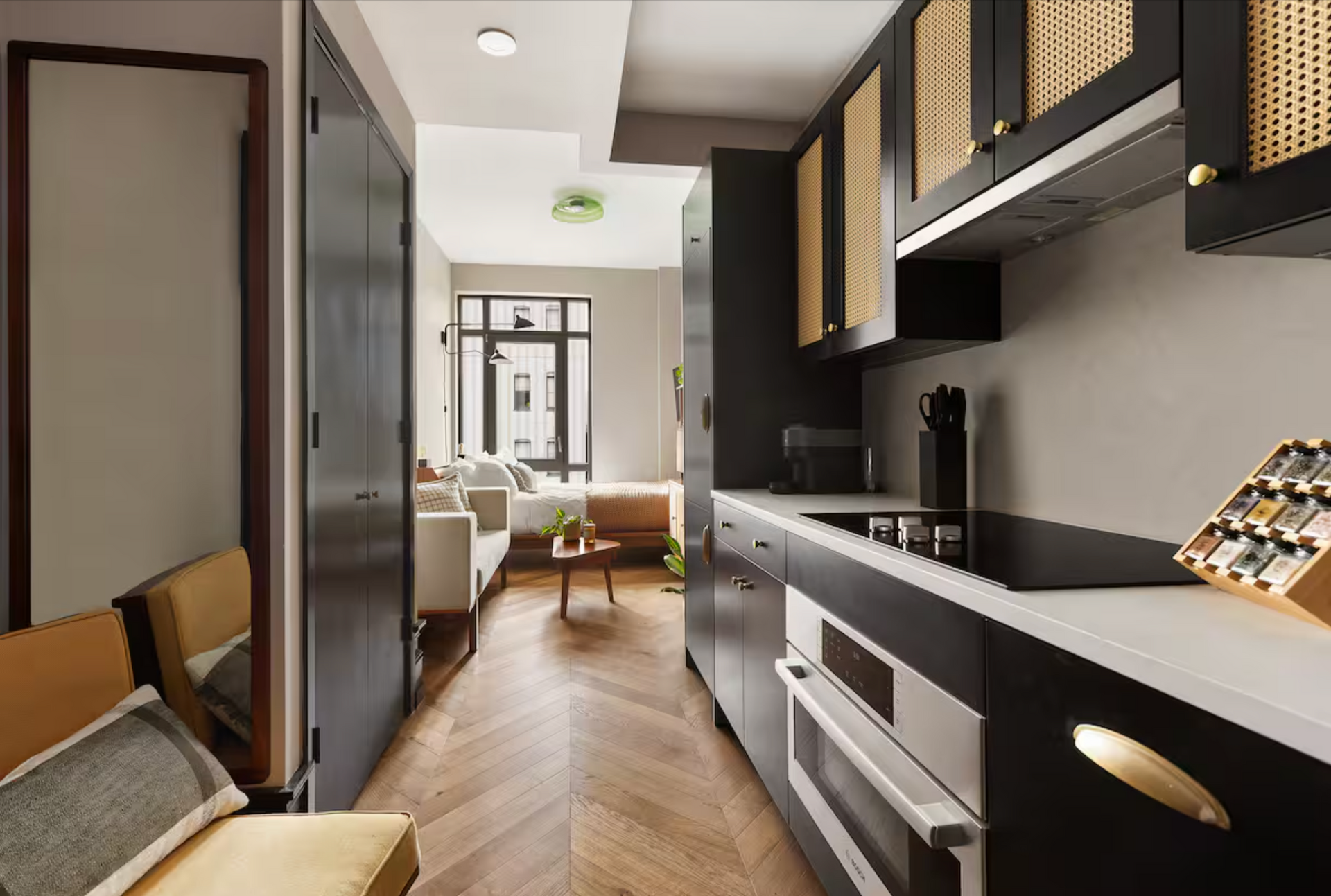 Small kitchen and hallway with dark cabinets, herringbone floor, and a living area in the background.