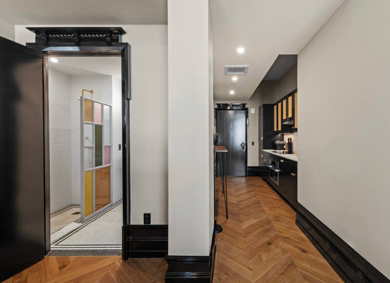 Hallway with herringbone wood floor, kitchen and bathroom visible. Black trim and accents.