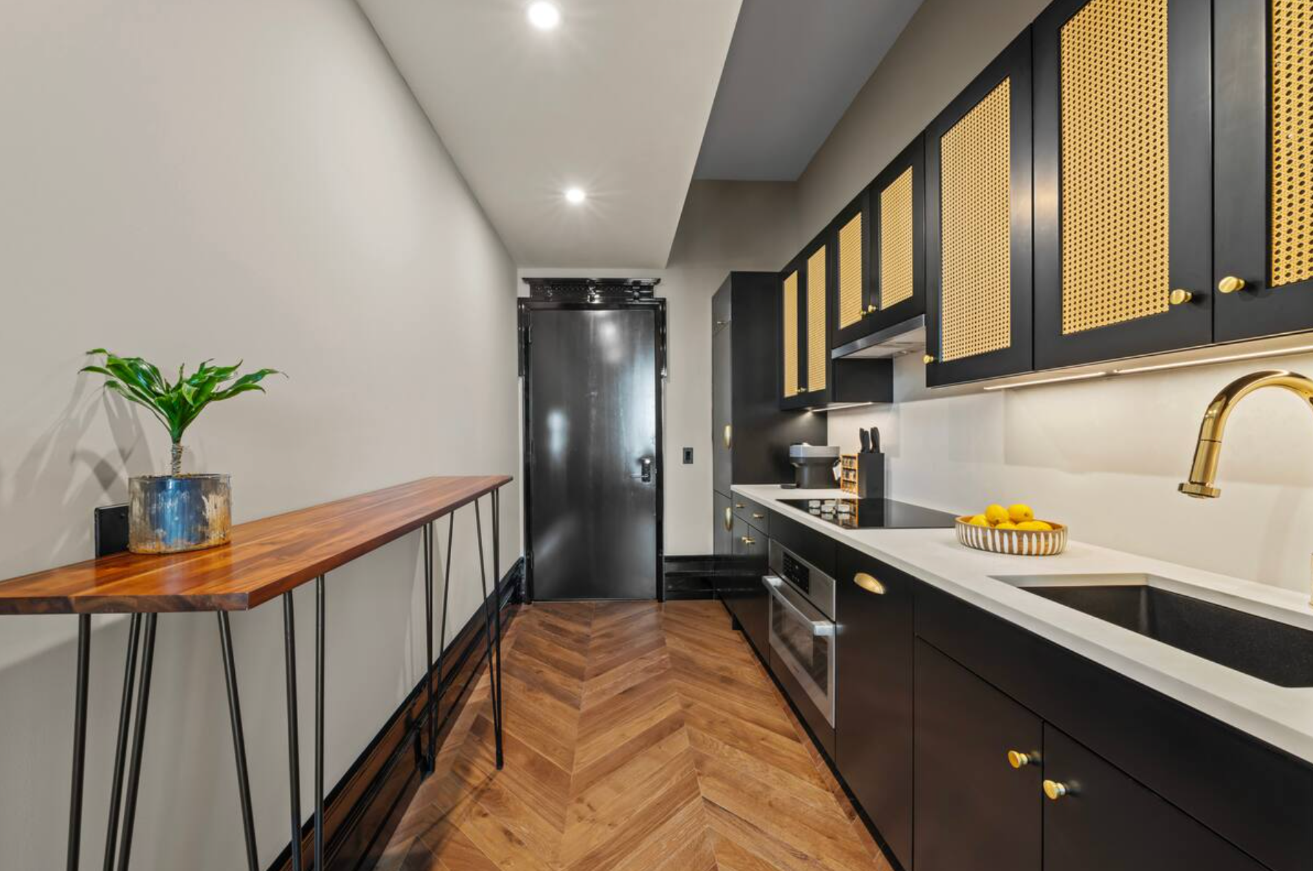 Narrow kitchen with black cabinets, gold accents, white countertop, and a wooden table.