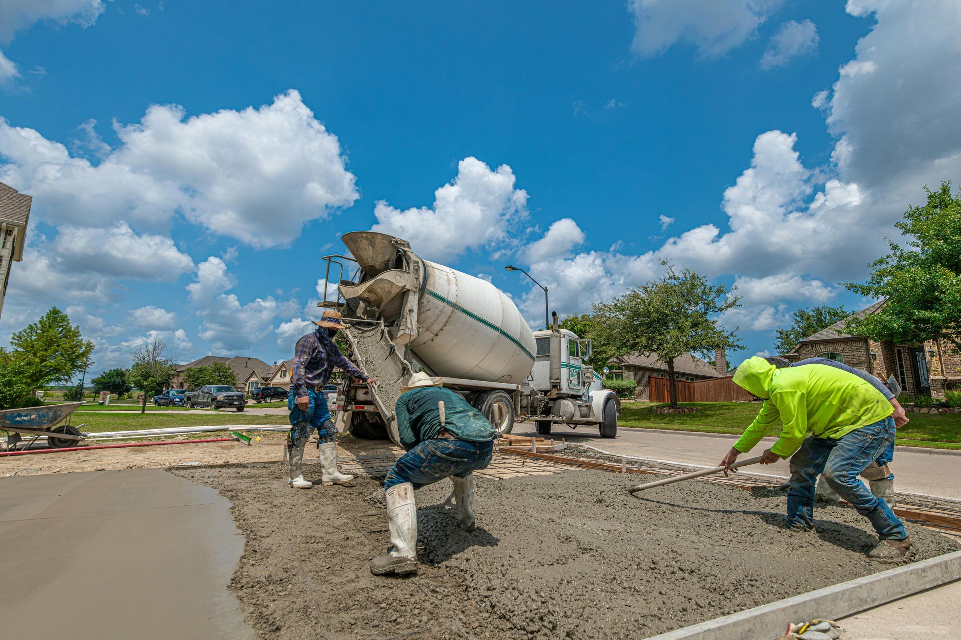 Construction workers use rakes to level fresh concrete poured from a mixer truck on a sunny day.