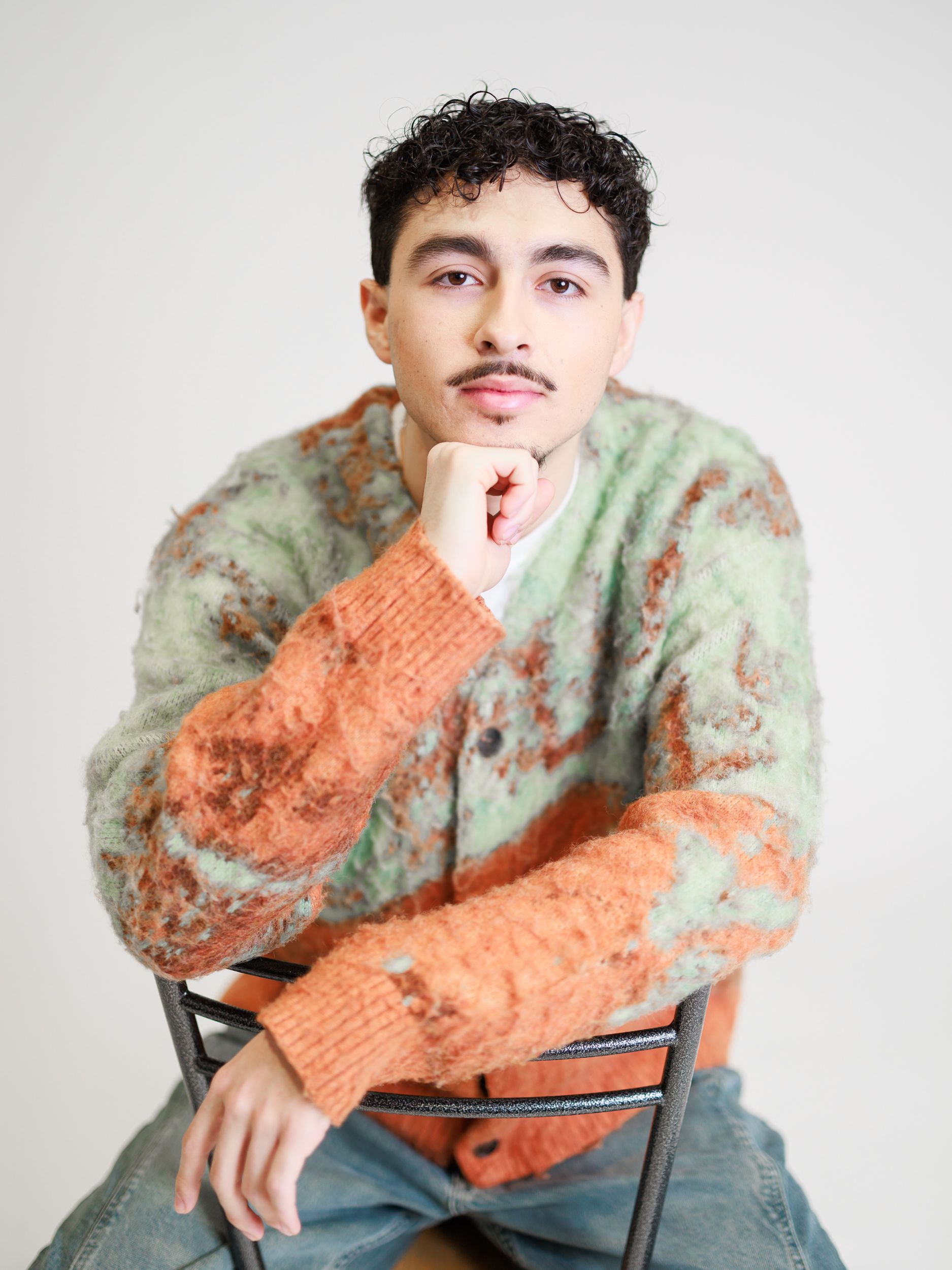 A young man is sitting on a chair with his hand on his chin.