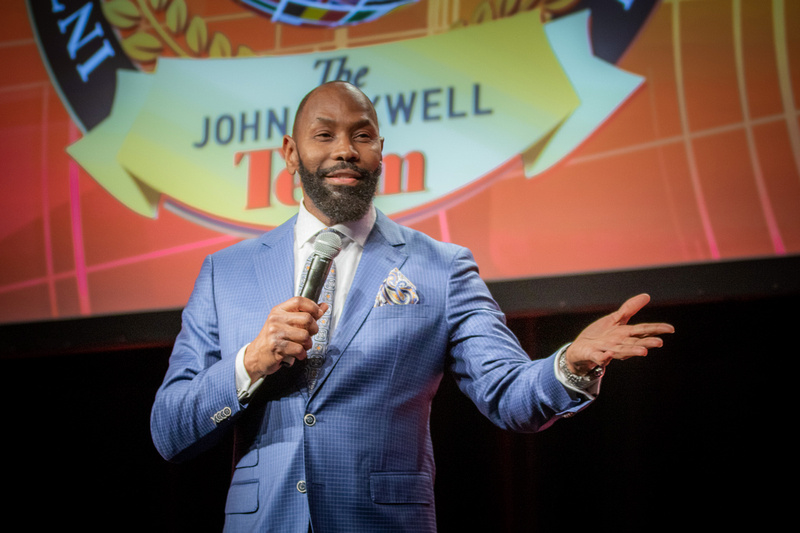 Man in blue suit speaking into a microphone on stage; 