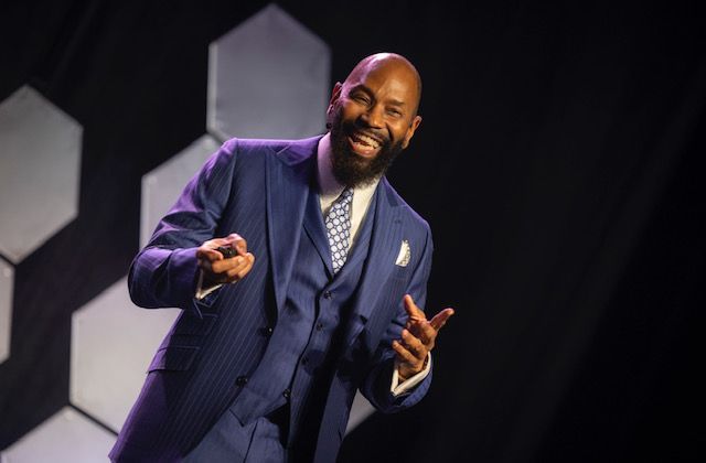 A Black man in a blue suit gestures on stage with a smile; geometric background.