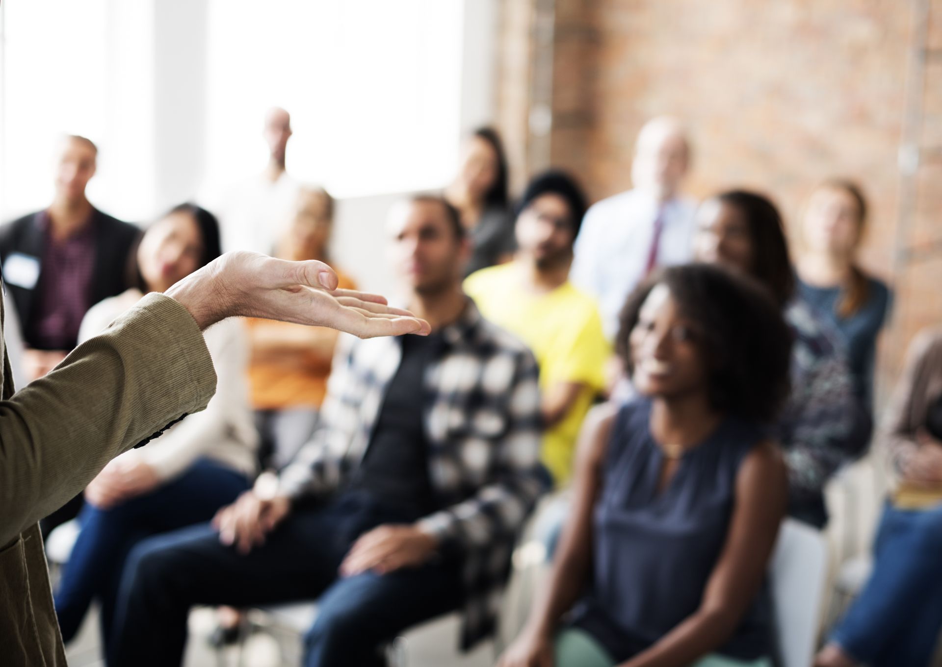 Person gestures while speaking to diverse audience seated in a room.