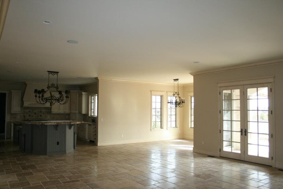 Empty, open-plan interior with kitchen island, two chandeliers, french doors, and beige tiled floors.