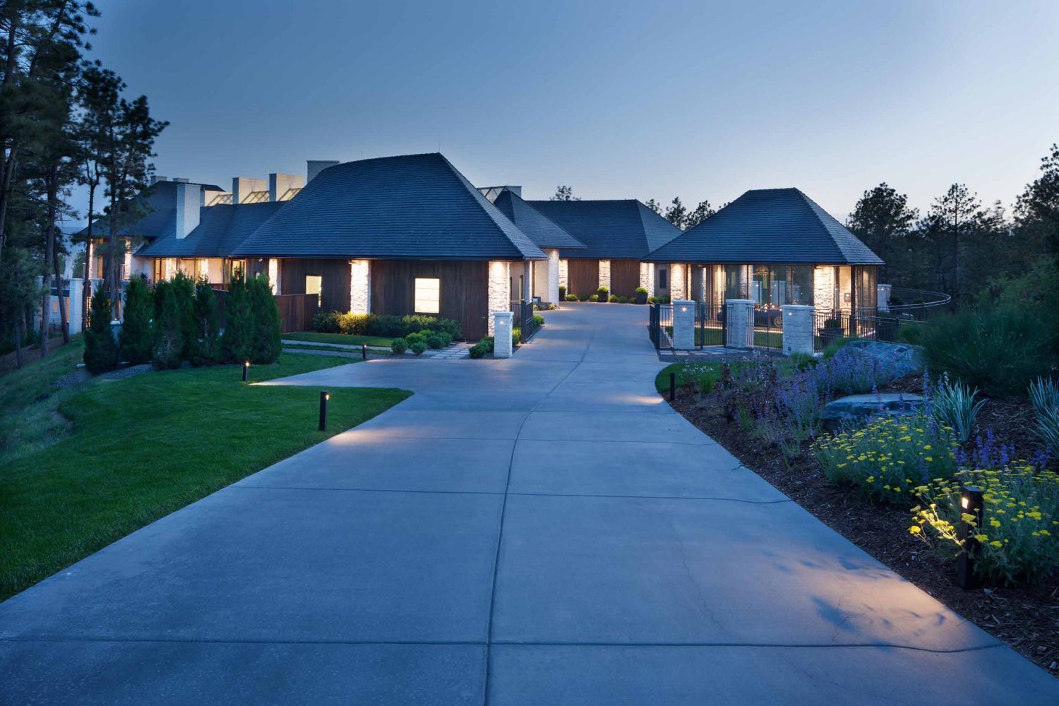 Long, gray driveway leading to a large, modern home with lit windows at dusk.