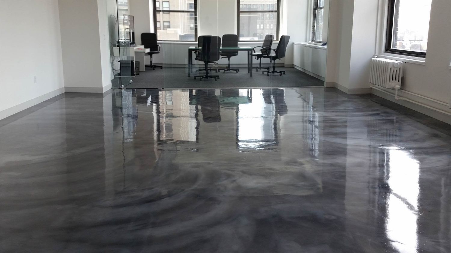 Shiny gray epoxy floor reflecting a bright office with furniture. Windows offer city views.